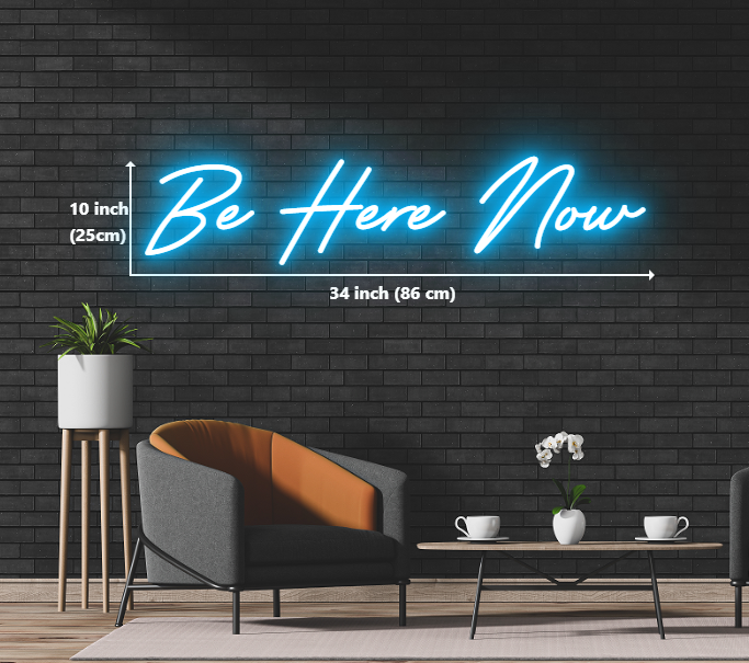 Neon sign with the words 'be here now' glowing in vibrant colors against a dark background.